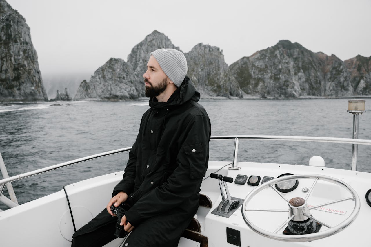 A man with a beard in a jacket sits on a boat steering through a rocky sea landscape.